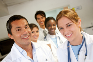 this a picture with fives doctors in a hallway. One is a male with brown hair smiling and there is a female with brown hair smiling in front of him on the left on the right is a lady with black hair  smiling in front of them is a male with brown hair smiling on the left, and on the right is a lady with redish blonde hair smiling